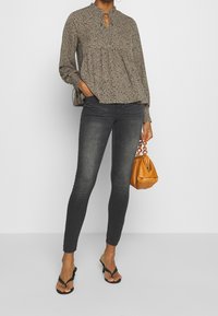 Blusa floreale color beige chiaro con scollatura a chiave, abbinata a jeans skinny scuri e infradito neri; tiene una borsa arancione.