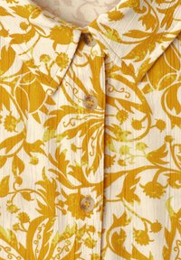 Chemise à col avec motif floral jaune sur tissu crème, avec des boutons beiges le long de la patte de boutonnage.