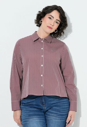 Kvinde med kort krøllet hår iført rød og hvid stribet skjorte og blå jeans, står foran en hvid baggrund.