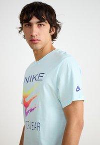 Šviesiai mėlyna medvilninė marškinėliai su spalvingu Nike logotipu, esančiu per krūtinę su gradientiniais atspalviais. Trumpomis rankovėmis ir prekės ženklo logotipas ant rankovės.