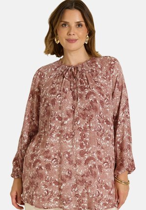 Mujer con blusa floral color malva de manga larga y holgada, escote fruncido, pendientes dorados y pulsera, mirando al frente y sonriendo.