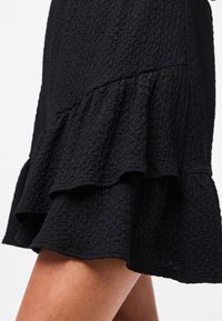 Gonna nera testurizzata con orlo a balze sovrapposte indossata da una persona, che mostra i dettagli e il movimento del tessuto.