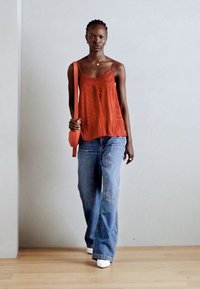 Haut sans manches rouge avec dentelle, associé à un jean large bleu clair. Le mannequin porte un petit sac à bandoulière orange et des chaussures blanches.
