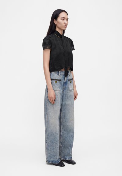 Femme portant un chemisier noir à manches courtes avec dentelle et un jean délavé à jambes larges avec poches avant, debout devant un fond blanc uni.