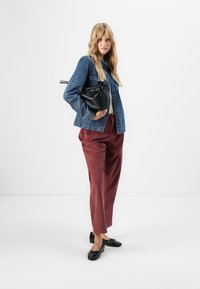 Woman wearing denim jacket, maroon pants, black flats, holding a black textured handbag, standing against a plain white background.