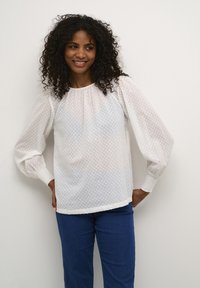 Vit blus med vita prickar, puffärmar, texturerat tyg och elastiska manschetter. I kombination med blå denimbyxor, bärs på ett avslappnat sätt.