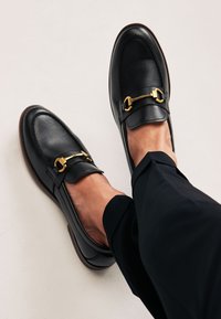 Les mocassins en cuir noir présentent un bout arrondi, une semelle plate et un accent en métal doré sur le devant, portés avec un pantalon sombre.