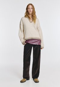 Bézs kötött pulóver V-nyakkivágással és bordázott szegéllyel, alatta bordó fényes felső. Fekete bőszárú nadrág, bézs bebújós cipővel párosítva.