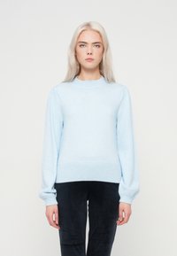 Gaiši zila džemperis ar apaļu kakla izgriezumu, strukturētām plecu krokām un ribām aprocēm un apakšmalai, valkāts virs tumšiem teksturētiem biksēm.