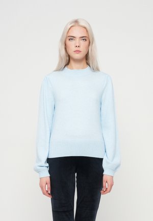 Pull en maille bleu clair avec un col rond, des plis structurés aux épaules et des poignets et un ourlet côtelés, porté sur un pantalon texturé sombre.