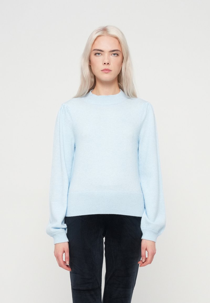 Gaiši zila džemperis ar apaļu kakla izgriezumu, strukturētām plecu krokām un ribām aprocēm un apakšmalai, valkāts virs tumšiem teksturētiem biksēm.