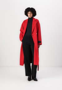 Roter Wollmantel mit langer Silhouette, Reverskragen und doppelter Knopfleiste, kombiniert mit schwarzem Rollkragenpullover und Hose.
