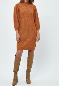 Femme portant une robe en maille côtelée couleur rouille avec des manches trois-quarts, assortie à des bottes en cuir marron clair montant jusqu'aux genoux.