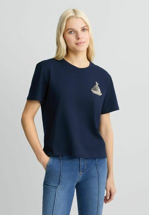 T-shirt à manches courtes bleu marine en coton doux avec un graphique de bateau sur la poitrine, doté d'un col rond et d'une coupe décontractée, associé à un jean bleu.