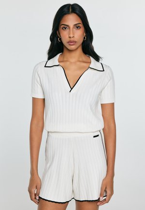 Sieviete ar gariem tumšiem matiem, tērpusies baltā ribotā īsroku krekla un atbilstošu šortu komplektā ar melnu apmali, stāvot pret vienkrāsainu fonu.