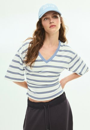 Jeune femme aux longs cheveux bruns portant une casquette Boston bleue, un t-shirt blanc et bleu à rayures et col en V, et un pantalon noir à cordon, mains sur les hanches.