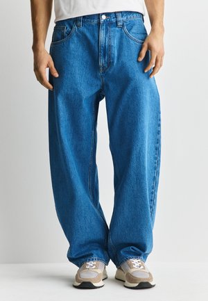 Person trägt lockere blaue Jeans und beige Sneakers, steht vor einem schlichten hellen Hintergrund.