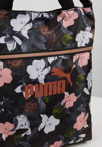 Musta kukkakuviollinen Puma-kangaskassi, jossa on vaaleanpunaisia ja valkoisia kukkia, vihreitä yksityiskohtia sekä etupuolella vetoketjullinen tasku, jossa on kuparinvärinen Puma-logo.