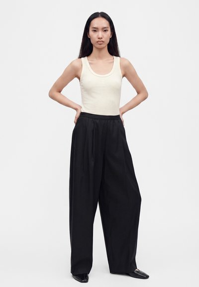Femme aux longs cheveux noirs portant un haut crème sans manches et un pantalon large noir ample, debout avec les mains sur les hanches.