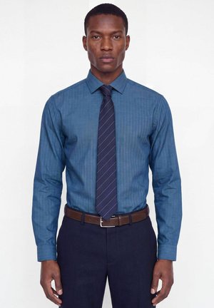 Homme portant une chemise bleue à fines rayures, une cravate rayée bleu foncé, un pantalon sombre et une ceinture en cuir marron, se tenant devant un fond uni.