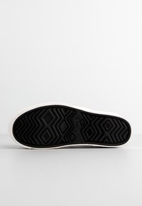 Semelle en caoutchouc noir avec motif géométrique de la bande de roulement et bord blanc, affichant le nom de la marque TOMS au centre.