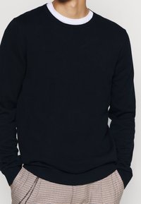 Marineblauer Strickpullover mit Rundhalsausschnitt und langen Ärmeln, ausgestattet mit einem gerippten Saum und Bündchen. Getragen über einem weißen Hemd und kombiniert mit karierten Hosen.