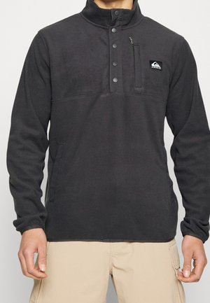 Mann trägt schwarzen Fleecepullover mit Druckknöpfen und Reißverschlusstasche auf der Brust, kombiniert mit beigen Cargoshorts.