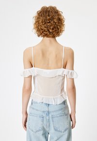 Weißes schulterfreies Oberteil aus durchscheinendem Stoff mit Rüschendetails, dünnen Trägern und einem Peplum-Saum, getragen mit hellblauen Jeans.