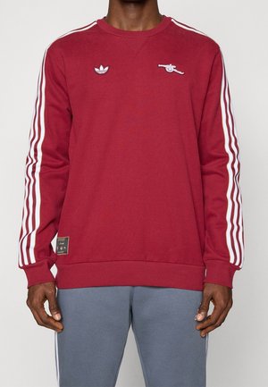 Mann trägt einen dunkelroten Sweatshirt mit weißen Streifen an den Ärmeln, zwei Brustlogos und graue Hose vor neutralem Hintergrund.