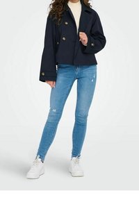 Chaqueta corta de doble botonadura en color navy con grandes botones, combinada con jeans ajustados desgastados de color azul claro y zapatillas blancas.