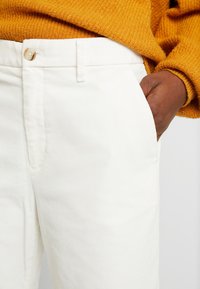 Vita bomullbyxor med en framficka och knappstängning i midjan, i kombination med en strukturerad senapsgul tröja.