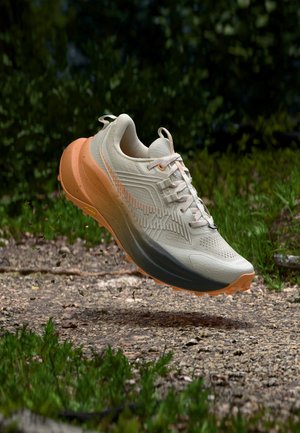 Chaussure de trail beige clair et orange flottant au-dessus d'un sentier forestier rocheux avec un feuillage vert en arrière-plan.