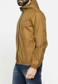 Giacca impermeabile marrone con cappuccio, chiusura con zip e tasche laterali. Texture liscia, materiale leggero e polsini elasticizzati.