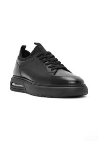 Zapatilla de cuero negra con una textura suave, que presenta una punta redondeada, frente con cordones, cuello acolchado y suela de goma gruesa con amortiguación de aire.