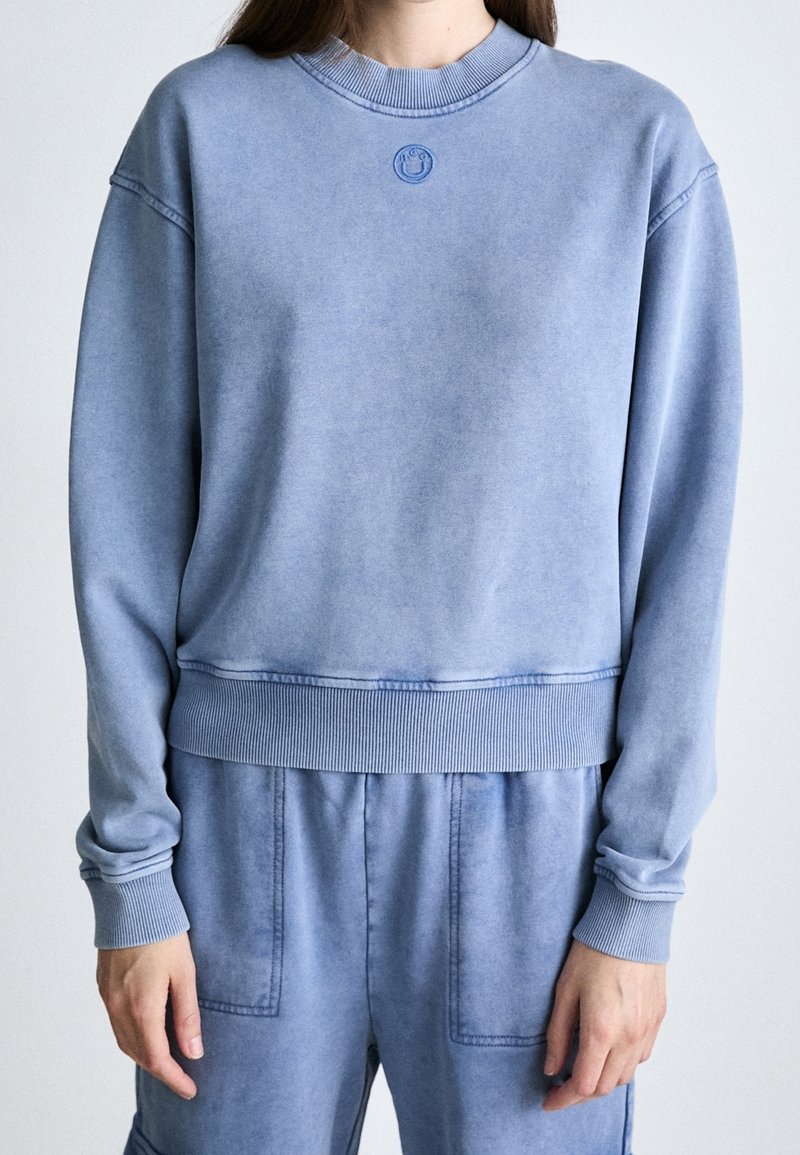 Personne portant un sweat-shirt à manches longues bleu clair et un pantalon assorti, debout avec les bras détendus devant un fond uni.