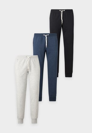 Uomo con t-shirt beige a maniche corte, pantaloni neri con coulisse e sneakers bianche, in piedi con le mani nelle tasche su sfondo bianco.
