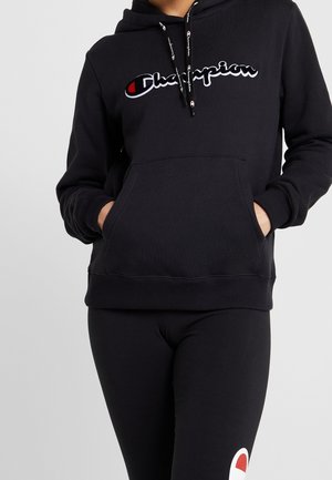 Person iført en sort Champion-hoodie med logo på brystet og matchende sorte leggings, hænderne i hoody-lommen, stående mod ensfarvet baggrund.