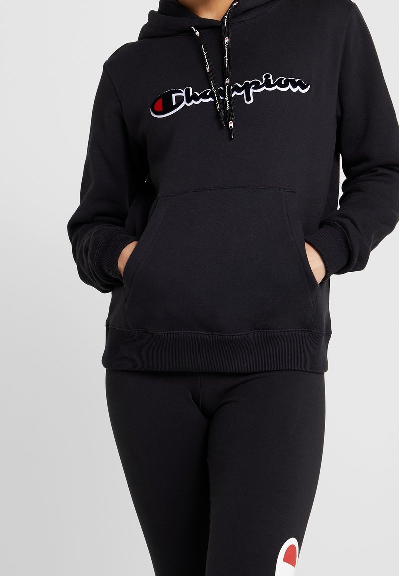 Personne portant un sweat à capuche noir Champion avec logo sur la poitrine et des leggings noirs assortis, mains dans la poche du sweat, debout devant un fond uni.