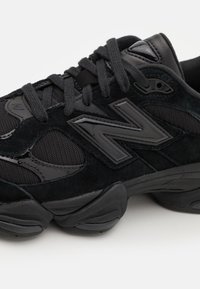 Gros plan d'une chaussure de sport noire avec une semelle épaisse, des panneaux en suède et en mesh, des lacets noirs et un grand logo stylisé « N » sur le côté.