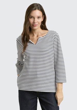 Jeune femme aux longs cheveux bruns, vêtue d'un haut à rayures blanches et bleues marine à manches longues et d'un jean foncé, debout devant un fond uni.