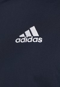 Logo Adidas blanc avec trois bandes diagonales brodées sur un tissu texturé bleu marine foncé.
