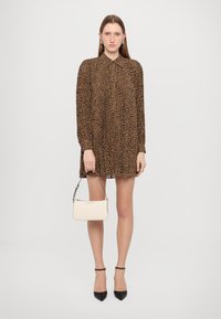Abito leopardato con maniche lunghe, colletto e design plissettato. Il modello tiene in mano una piccola borsa bianca e indossa tacchi a punta neri.