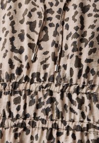 Kangastuskuvioinen kangas vaaleassa beige-värissä, jossa on tummia ruskeita pilkkuja. Kankaassa on rypytetty yksityiskohta, nyörit metallisilla päillä ja pehmeä rakenne.