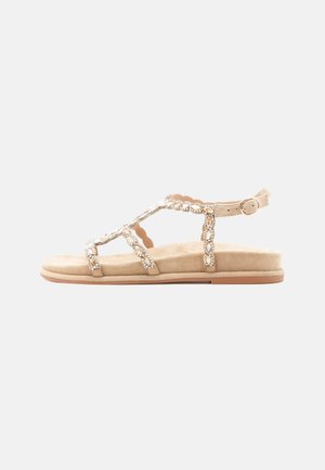 Beige platte sandaal met versierde T-riem, enkelriempje met gesp en gevoerde zool tegen een effen lichte achtergrond.