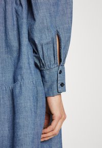 Vestido de denim com mangas longas, pregueadas, com punhos de botão e fendas laterais. O tecido tem um tom azul claro suave e um acabamento texturizado.