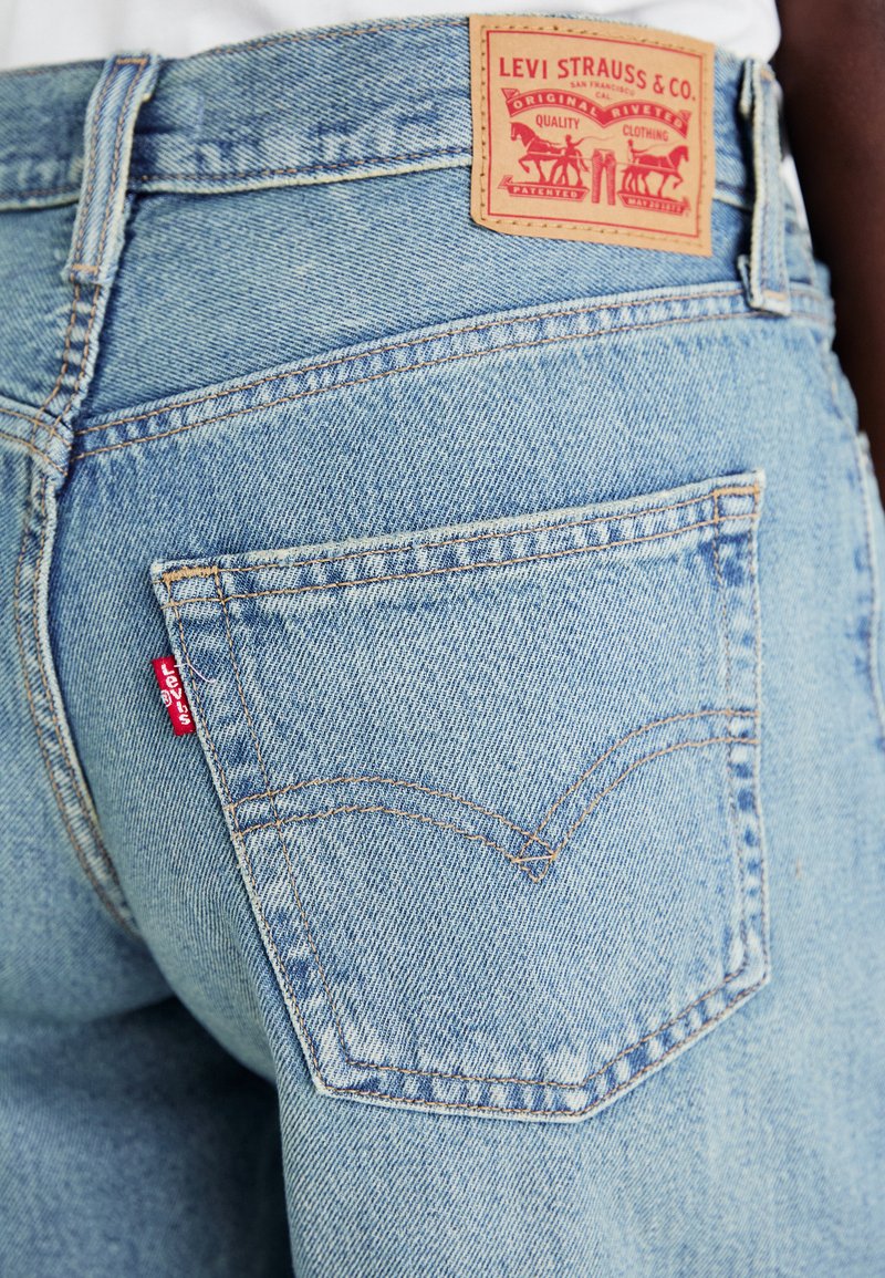 Jean en denim bleu clair avec une poche arrière visible, une étiquette rouge Levi's et une patch en cuir marron avec un logo de cheval et les détails de la marque.