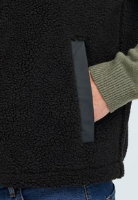 Giacca in pile nera con superficie testurizzata, maniche verdi a contrasto e una tasca laterale nascosta con un elegante pannello nero.