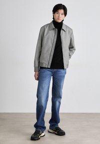 Grå teksturert jakke med ribbede mansjetter over en svart høyhalset genser, kombinert med blå rett bukser og mønstrede sneakers. Minimalistisk design.