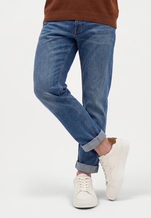 Uomo con jeans blu risvoltati e sneakers bianche, in piedi con una gamba incrociata sull’altra.