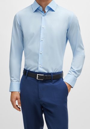 Mann trägt ein hellblaues, geknöpftes Hemd, steckt es in eine dunkelblaue Anzughose mit schwarzem Gürtel, Hand in der Tasche.